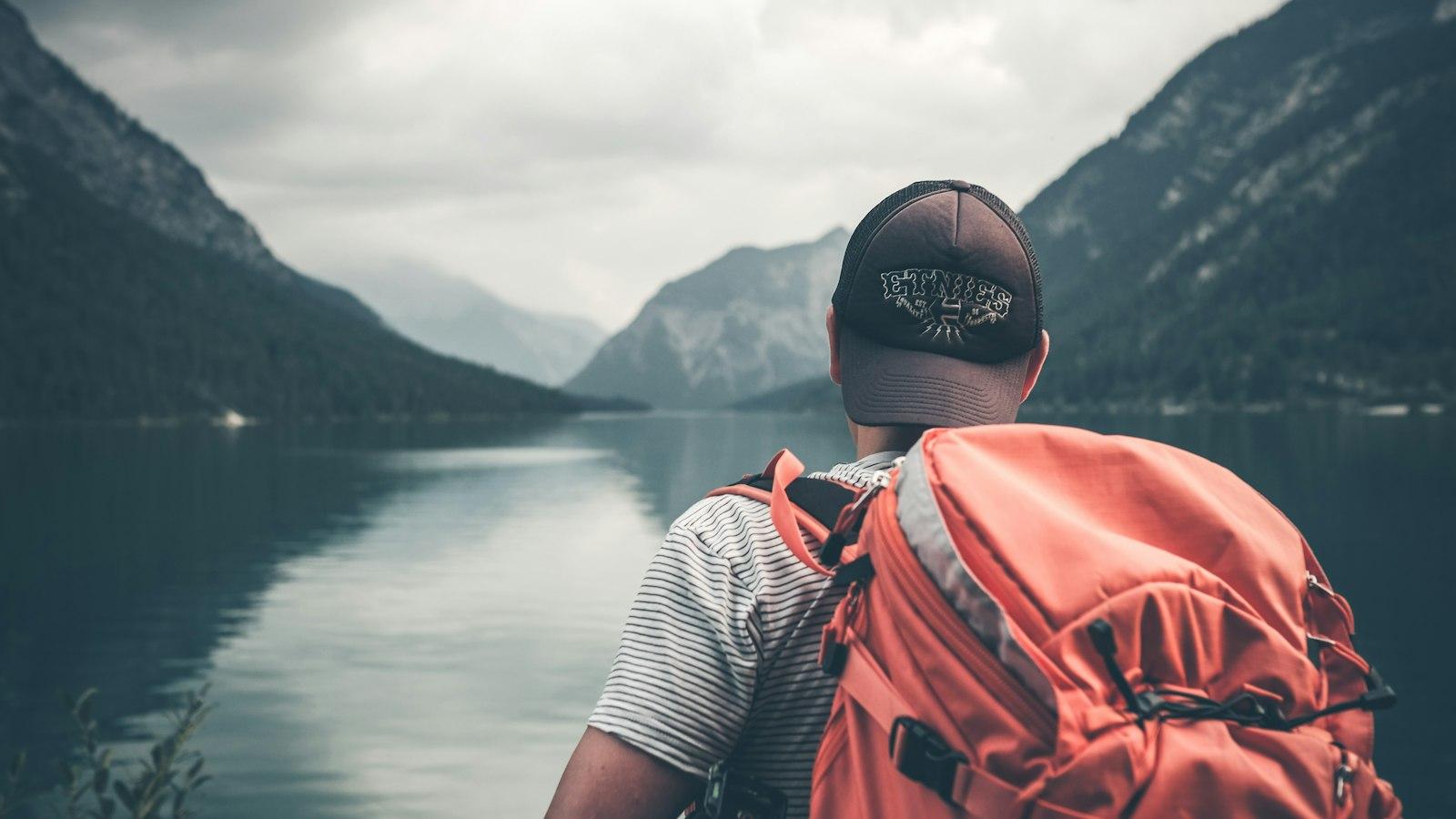 A traveler with a red backpack looking out over a mountain lake