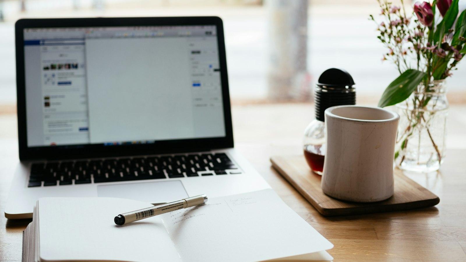 A laptop on a wooden table next to an open notebook, a coffee mug, and fresh flowers